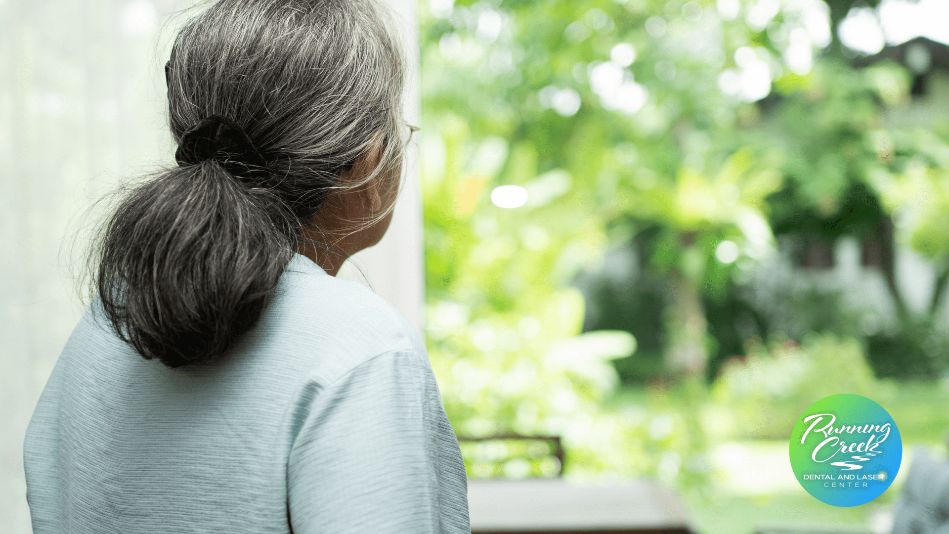 An older woman is sitting in front of a window looking out.